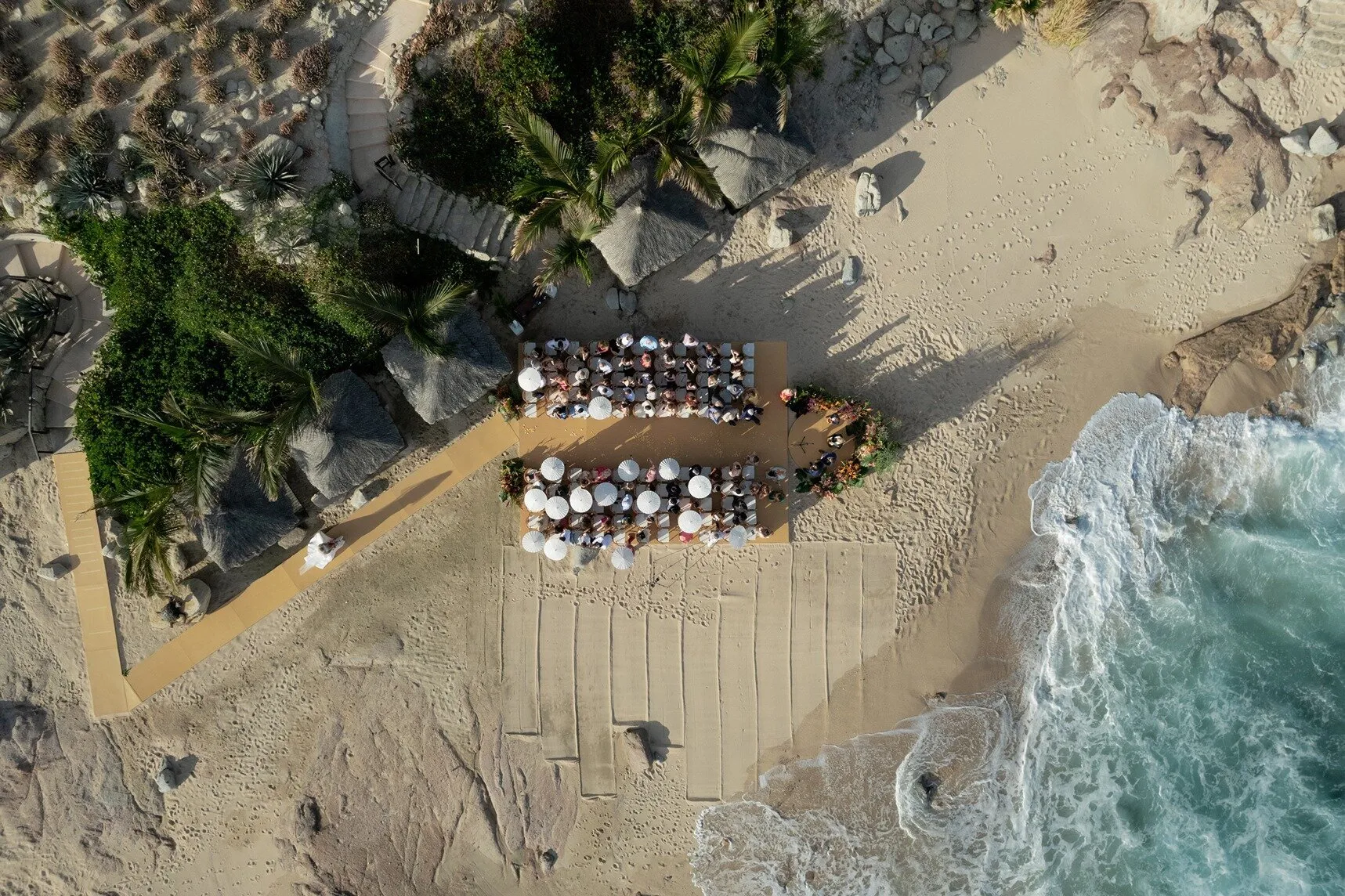 A beach ceremony seen from above — rows of chairs on the sand, the surf breaking a few metres away